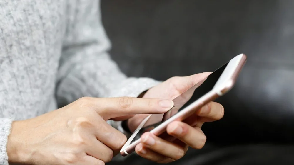 A person wearing a gray sweater is using a smartphone. One hand holds the phone while the other hand taps the screen.