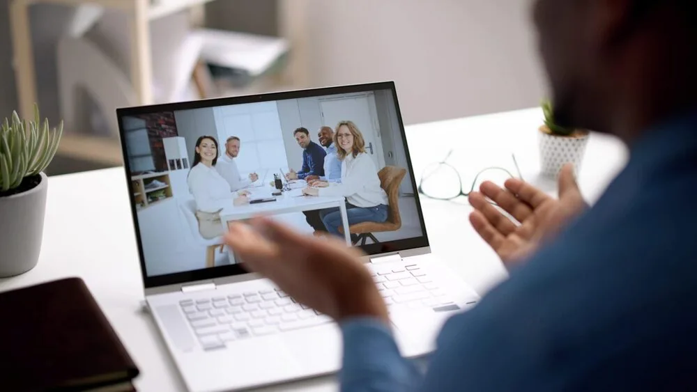 A person participating in a virtual meeting, viewing a laptop screen displaying five people sitting at a conference table. The scene is set in a modern, bright office with notebooks and plants nearby.