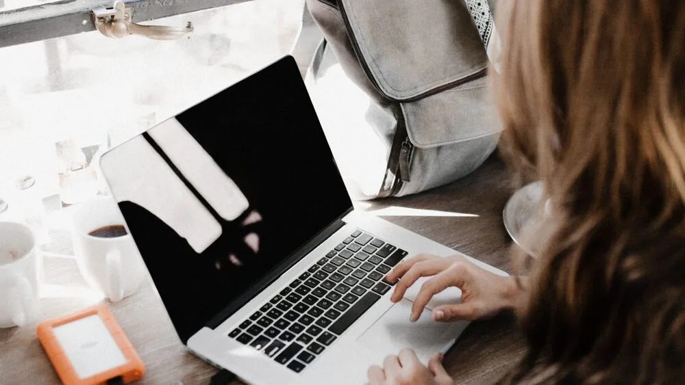 A person using a laptop at a wooden table, surrounded by a cup of coffee, an orange external hard drive, and a gray backpack. 