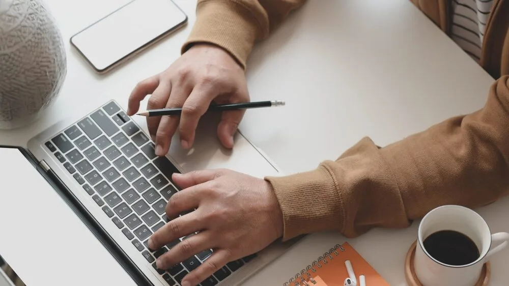 A person wearing a brown jacket types on a laptop while holding a pen. Nearby, there is a smartphone, a patterned vase, an orange notebook with a pen, and a cup of black coffee on the desk.