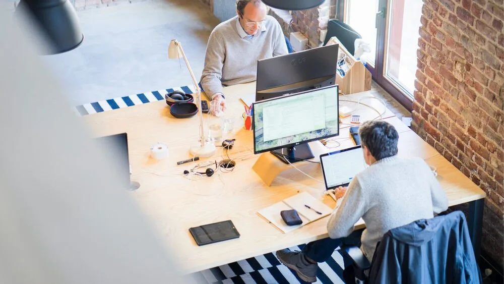 Two people working at a wooden desk with laptops in a modern office. 