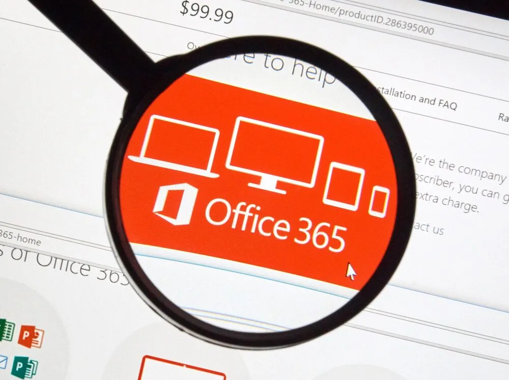 A magnifying glass hovers over a screen displaying the Microsoft Office 365 logo, which features icons of devices such as a desktop, laptop, tablet, and smartphone. 