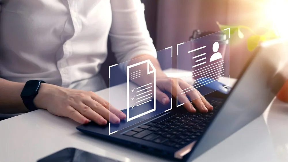 A person types on a laptop, with digital icons of a document and user profile hovering above the keyboard.