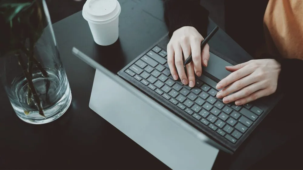 A person is typing on a detachable keyboard connected to a tablet in a dark workspace.