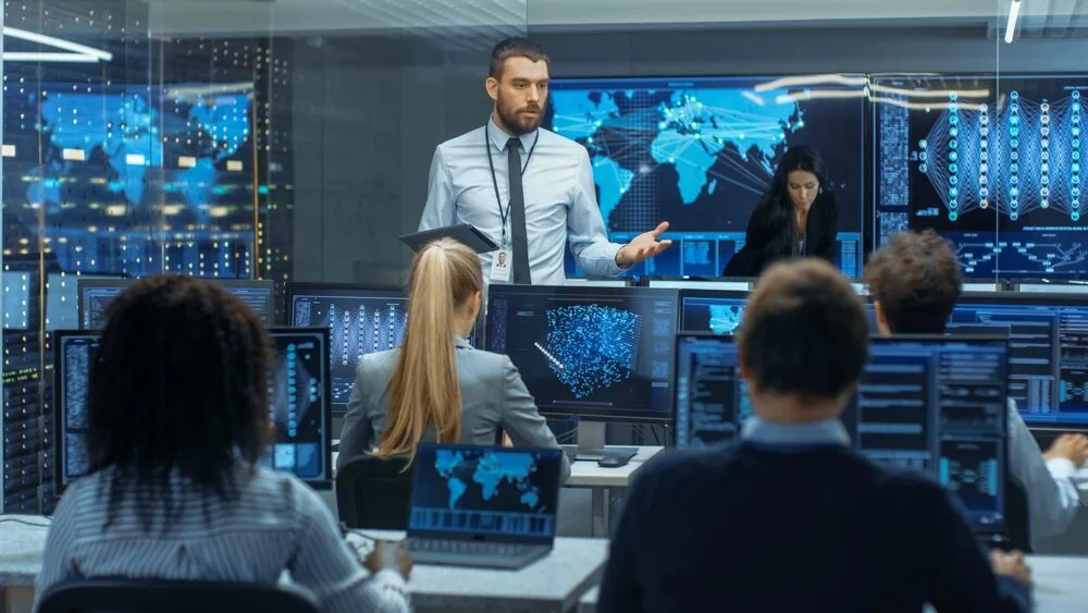 A man in a white shirt and tie stands in front of several computer monitors in a high-tech office, leading a meeting. 