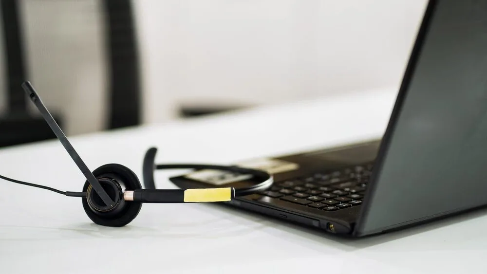 A black headset with a microphone is placed next to a black laptop on a white desk.