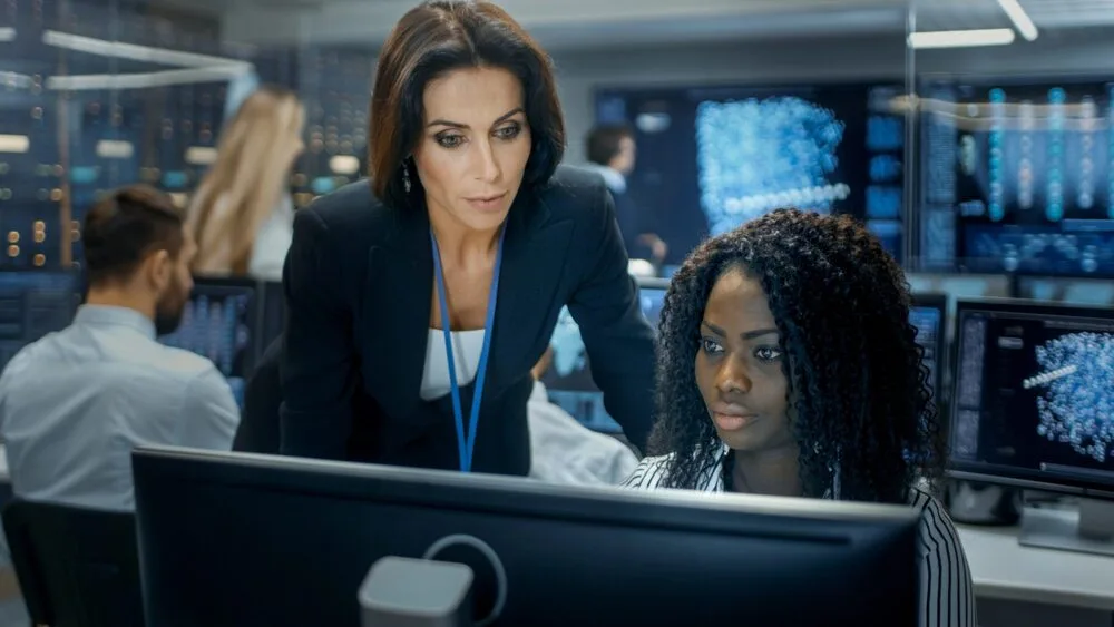 Two women are working in a busy office with multiple computer monitors displaying data. 