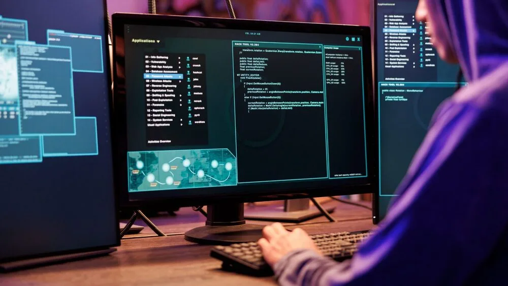 A person in a hoodie works at a desk with multiple computer monitors displaying code and application lists.