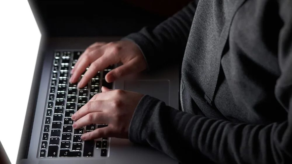 A person is typing on a laptop keyboard. The screen is bright, illuminating their hands in a dimly lit setting.