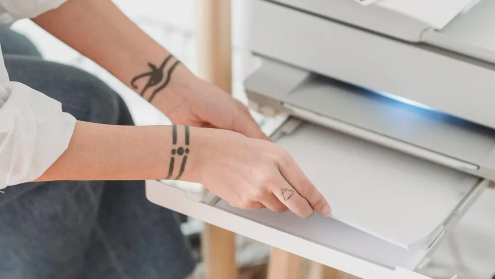 A person with geometric tattoos on their arm is placing paper into a printer, preparing it for printing. 