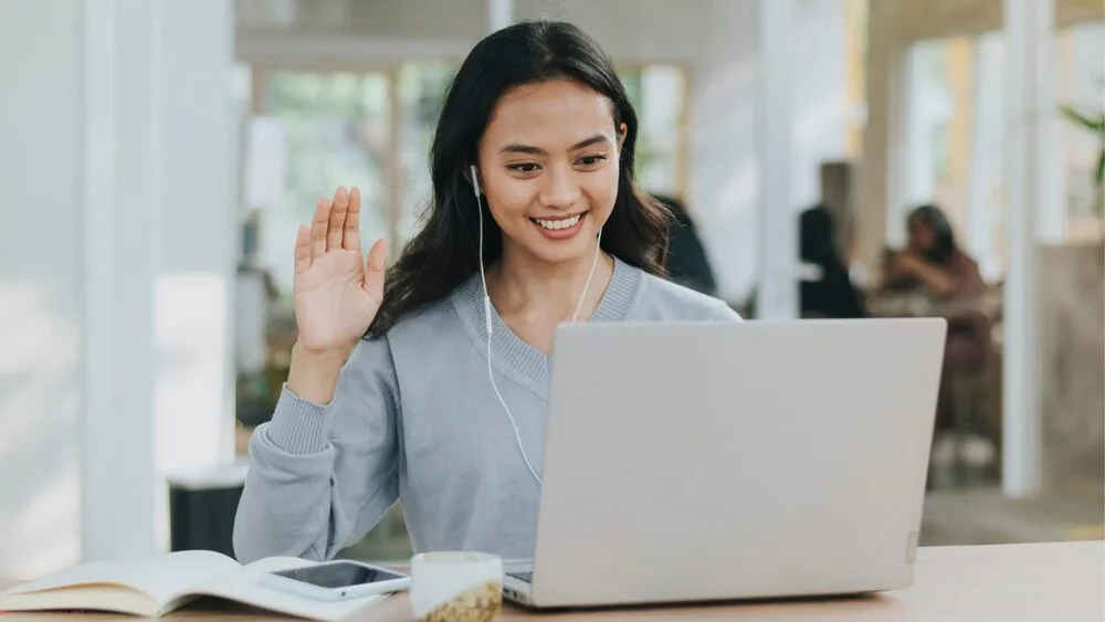 A person with long hair is sitting at a desk, smiling and waving while on a video call.