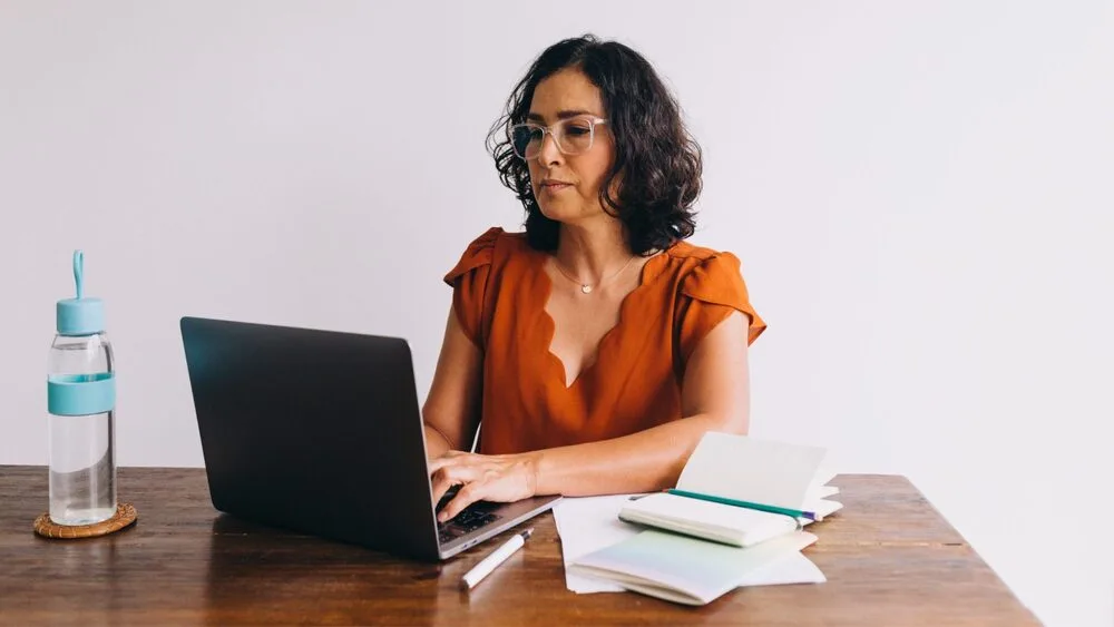 A woman with glasses and curly hair works on a laptop at a wooden table. 