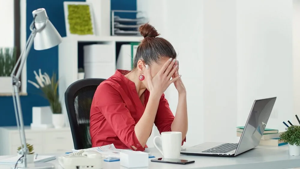 A woman in a red shirt sits at a desk with her head in her hands, appearing stressed.