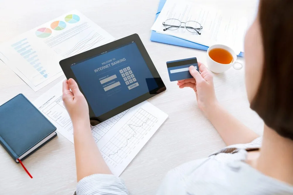 A person holds a tablet displaying an internet banking login page and a credit card.