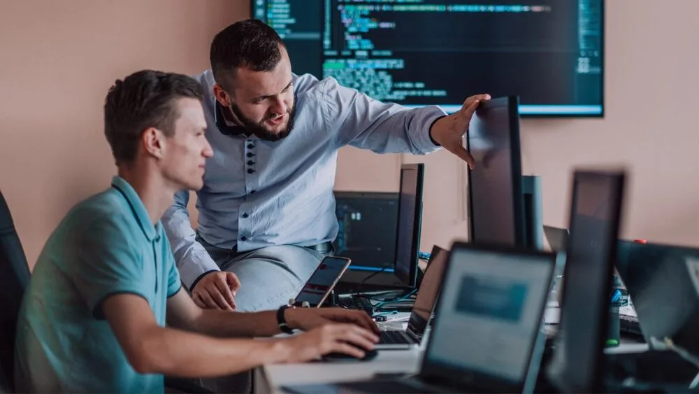 Two men in an office environment work at a desk with multiple computer monitors.