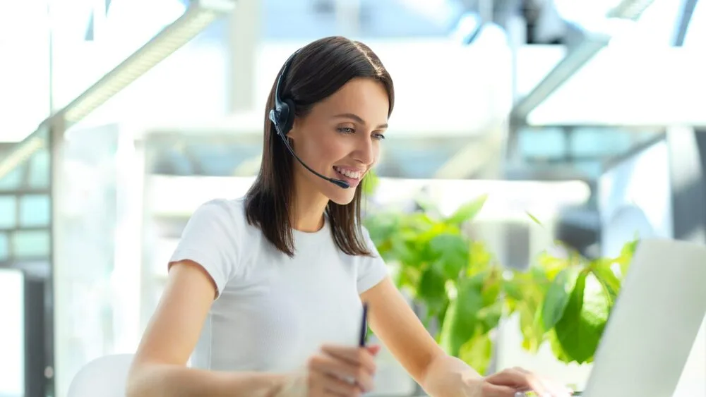 A woman with long brown hair is smiling and working at a computer while wearing a headset. 