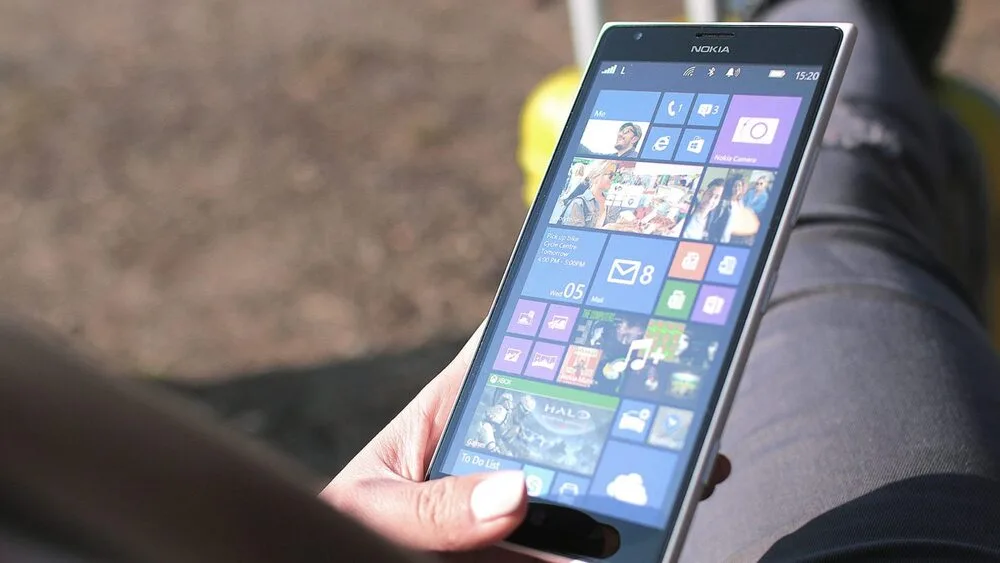 A person holding a Nokia smartphone displaying the Windows Phone home screen with various colorful tiles.