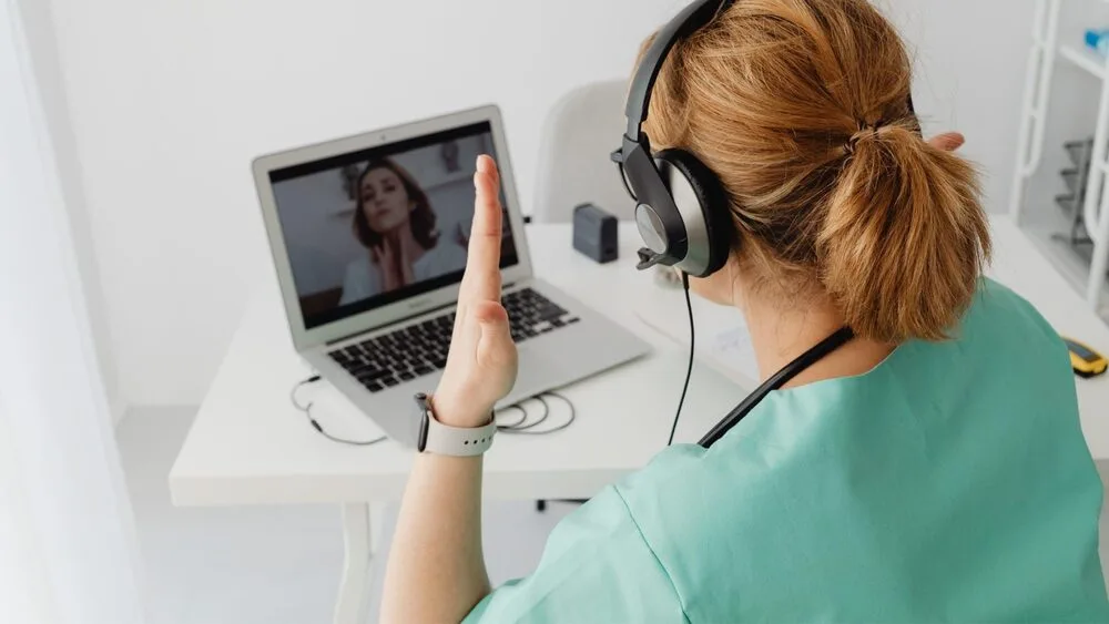 A woman wearing headphones sits at a desk, engaged in a video call on a laptop. 