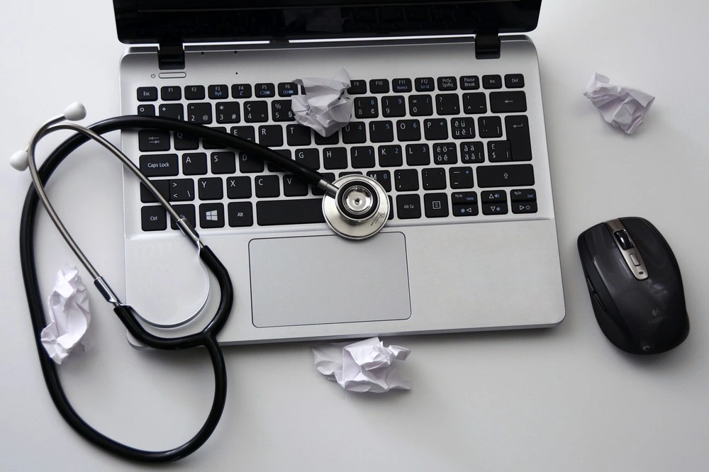 An open laptop with a stethoscope placed on the keyboard, surrounded by crumpled paper balls. A wireless mouse sits to the right of the laptop on a white surface.