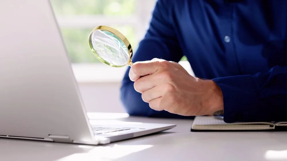 A person in a blue shirt holds a magnifying glass up to the screen of an open laptop. 