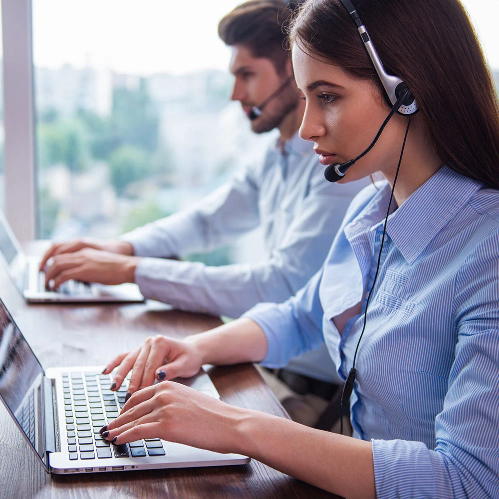 Two customer service representatives wearing headsets work on laptops at a wooden desk.