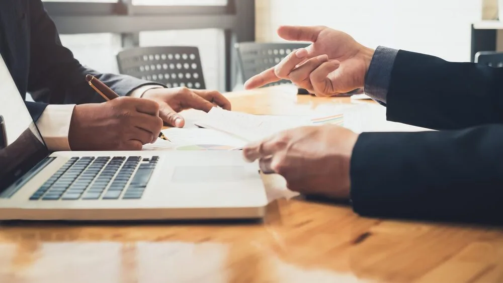 Two people in business attire are sitting at a desk with a laptop in front of them.