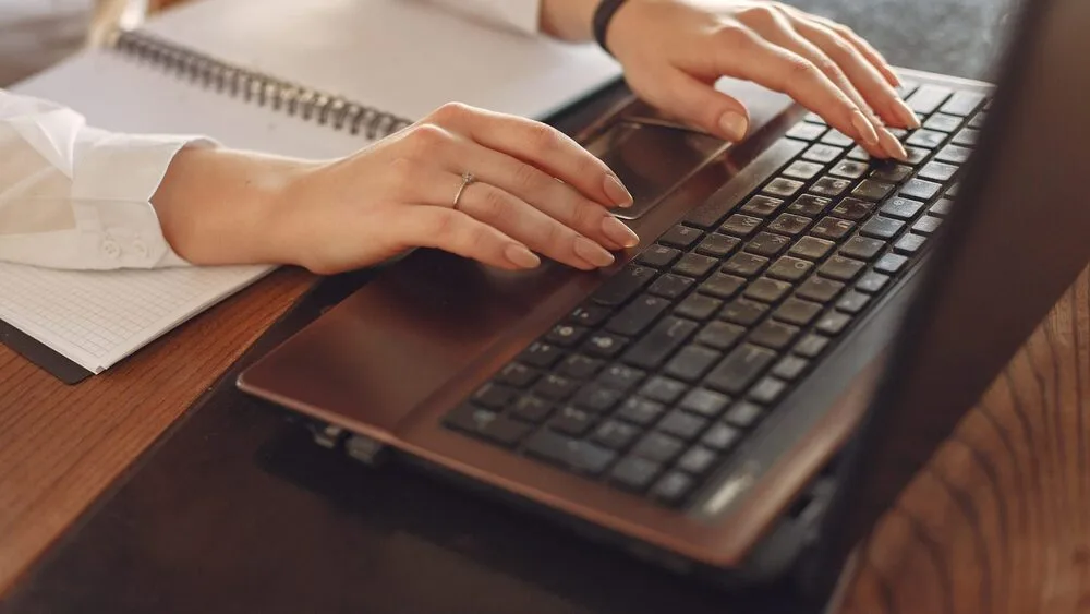 A person types on a laptop while a notebook lies open beside them on a wooden table. 