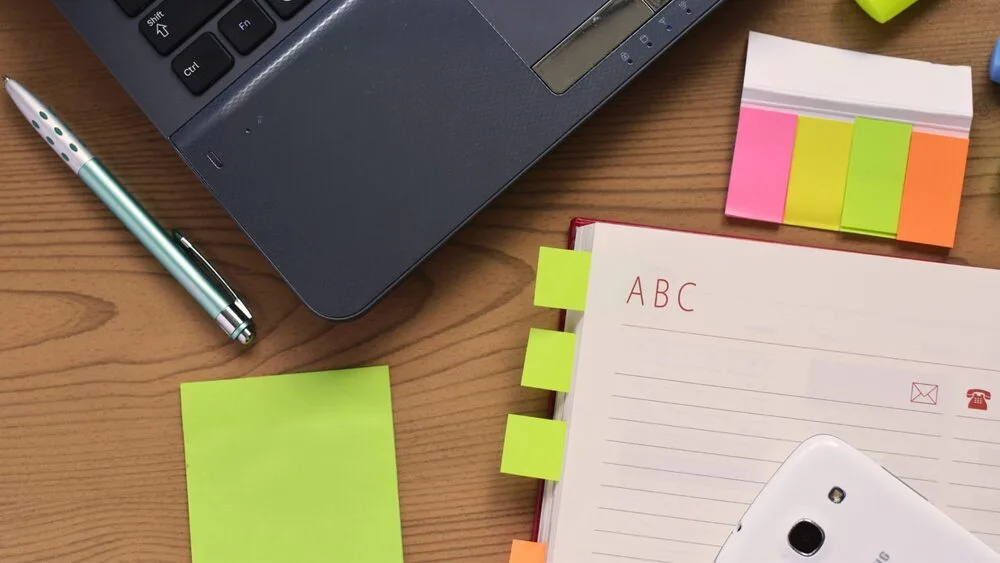 A wooden desk with a laptop, multicolored sticky notes, a pen, and an open notebook with tabbed sections labeled with letters.