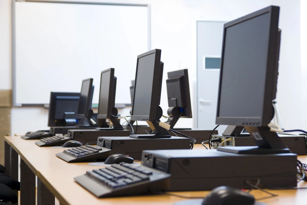 A classroom with several desktop computers placed on wooden tables in a row, each with a monitor, keyboard, and mouse. A whiteboard is visible in the background. The room appears organized and ready for use.