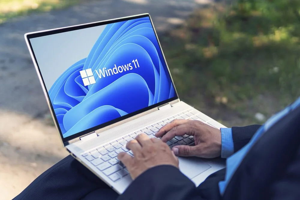 A person is using a white laptop outdoors with Windows 11 displayed on the screen. 