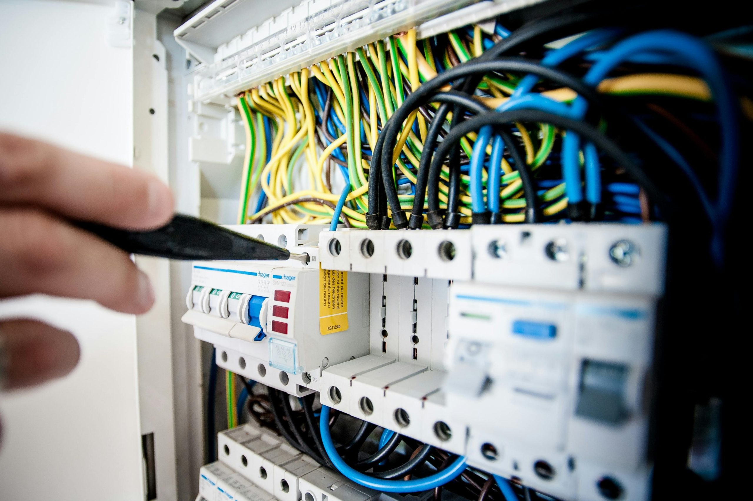 A hand holds a tool near an electrical circuit breaker panel filled with tangled, colorful wires. The circuit breakers and switches are visible, displaying various labels and markings.