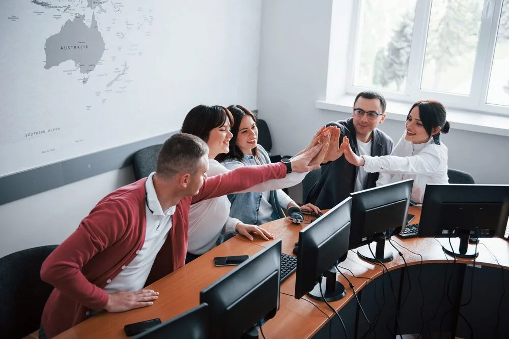 A group of six people, seated at a curved desk with computers, are enthusiastically high-fiving each other.