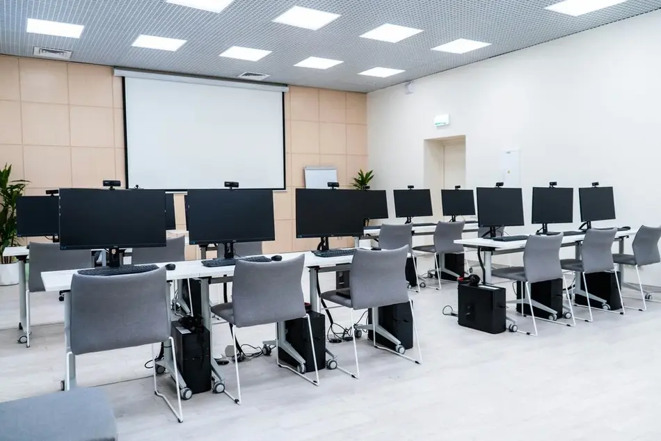 A modern computer lab with multiple workstations arranged in two rows, each equipped with a monitor, keyboard, and mouse. Gray chairs are placed at each station, and a large projector screen is mounted on the front wall. The room is well-lit and organized.