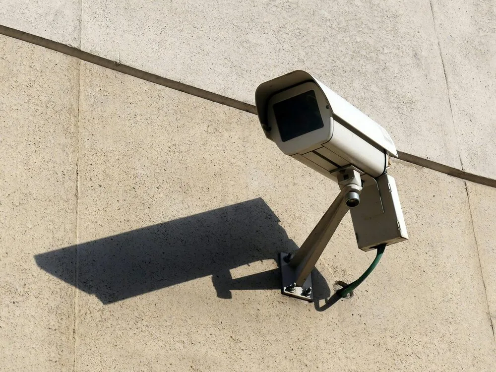 A security camera mounted on a beige concrete wall casts a shadow. 