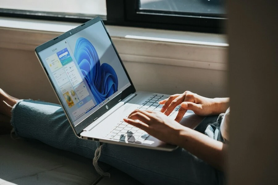 A person is sitting on a windowsill, typing on a silver laptop that displays the Windows 11 operating system on the screen. Sunlight streams through the window, illuminating the scene. The person is wearing casual clothing, including jeans.