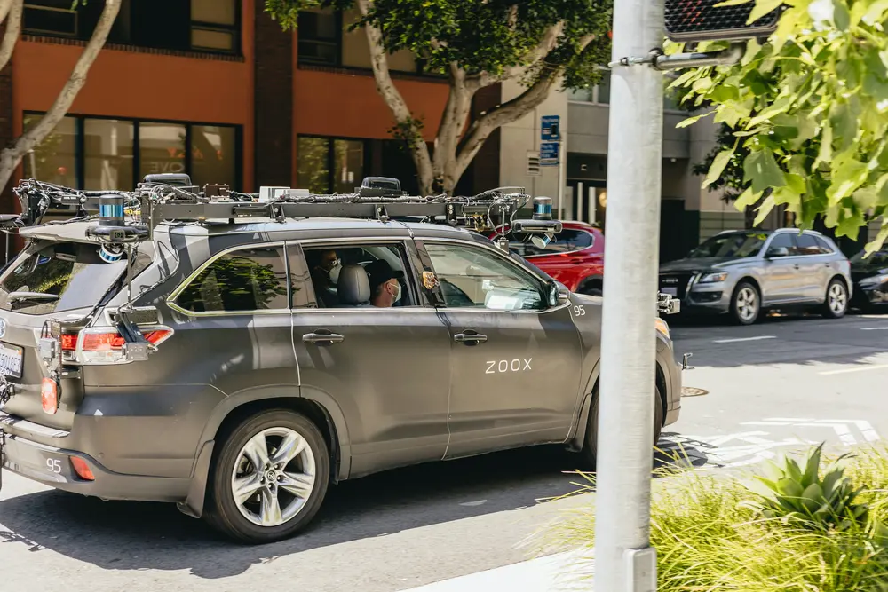 A Zoox autonomous vehicle is driving on a sunny street lined with trees and parked cars. 