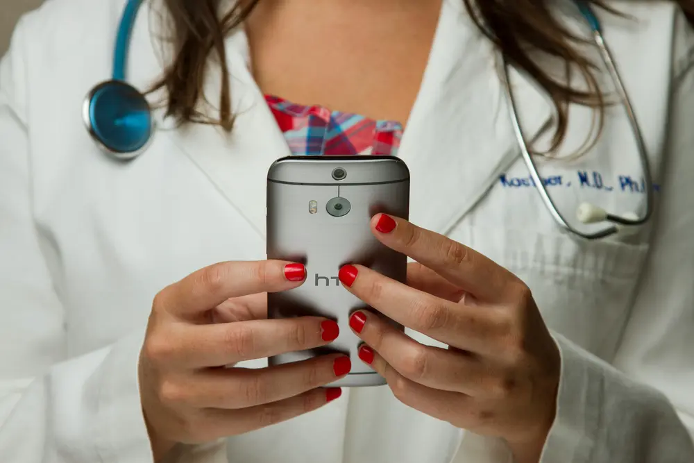 A doctor in a white coat with a stethoscope around their neck holds a smartphone. 