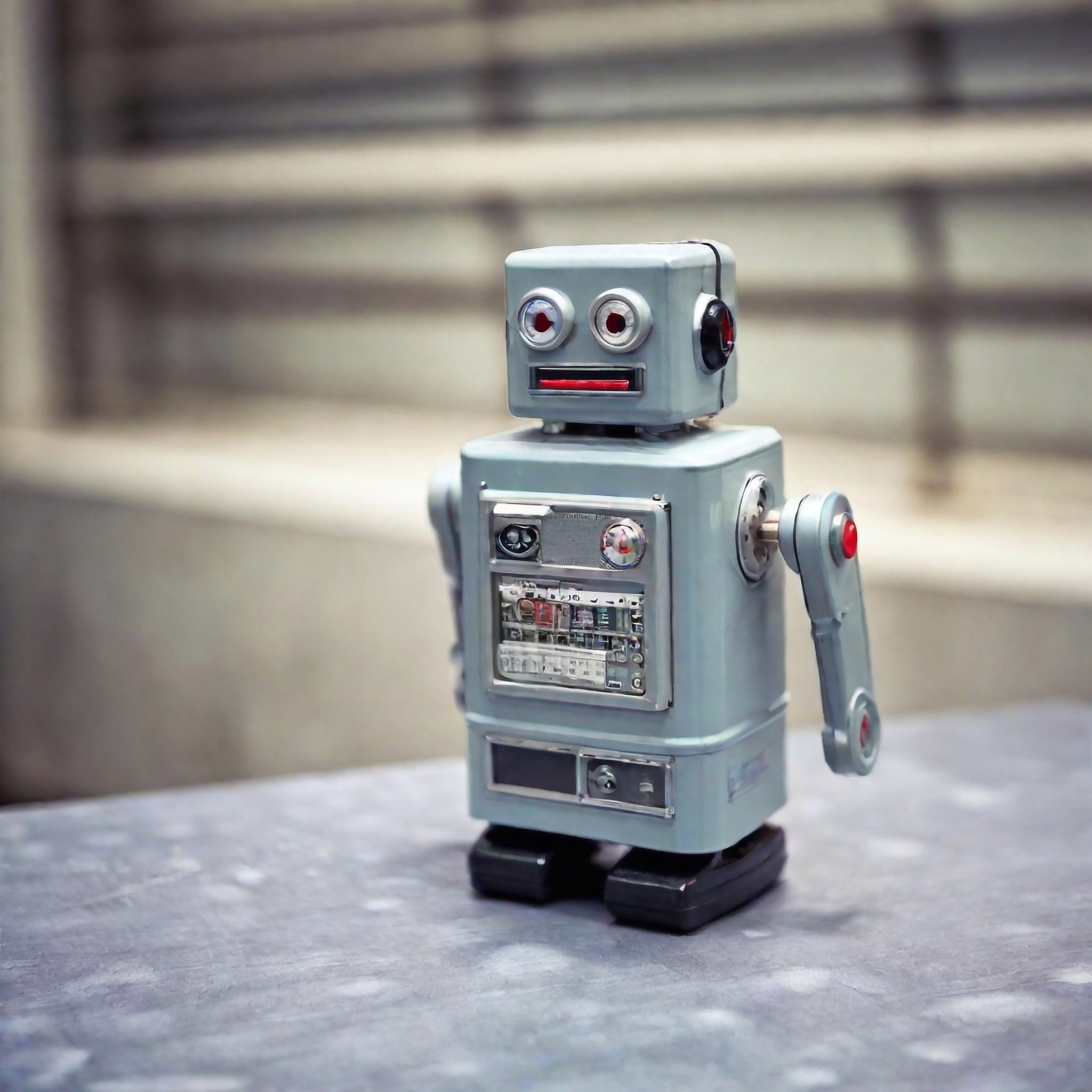 A vintage toy robot with a shiny metallic body, rectangular shape, and square head stands on a gray surface. It has circular eyes, a red mouth, and visible gears on its chest. The background is blurred, featuring horizontal lines.