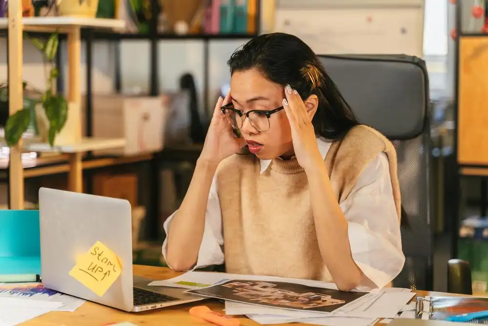 A person sitting at a desk looks stressed while holding their temples. They are in front of a laptop, with a sticky note reading "start up!" on it. Papers are spread across the desk, and a shelf with books and plants is visible in the background.