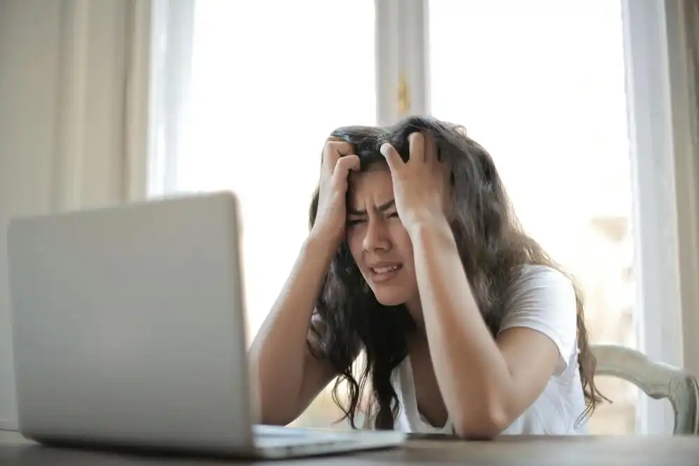 A woman in a white shirt sits in front of a laptop with her hands in her hair, looking frustrated. A window in the background lets in natural light.