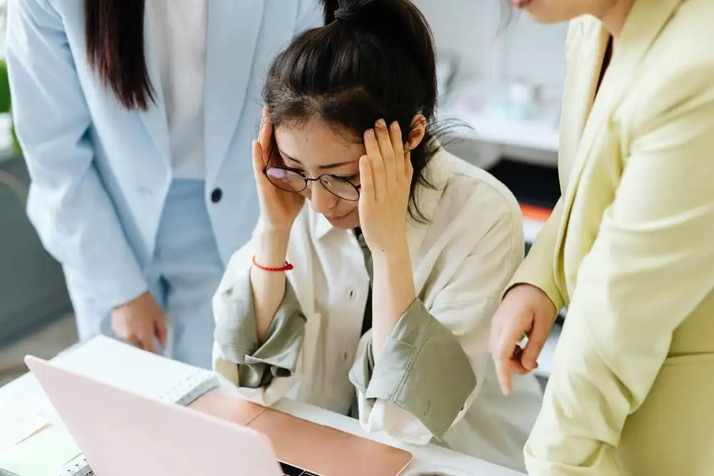 A woman sitting at a desk looks stressed while holding her head, surrounded by two colleagues. They are standing next to her, showing concern. An open laptop and paperwork are visible on the desk.