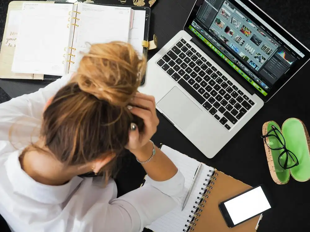 A woman sits at a desk with her head in her hands, surrounded by an open laptop, notebooks, and a smartphone. The desk has a cluttered appearance, and the laptop screen displays a photo editing software with various images.