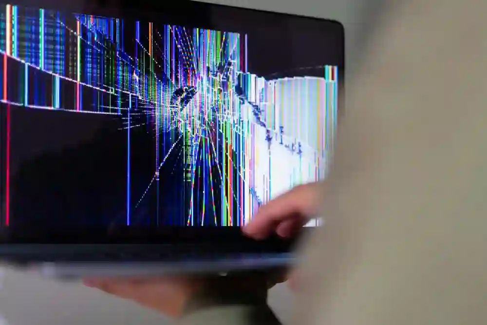 A person holds a laptop displaying a broken screen with colorful vertical and diagonal lines and a large black crack in the center, indicating severe damage. The background is blurred.