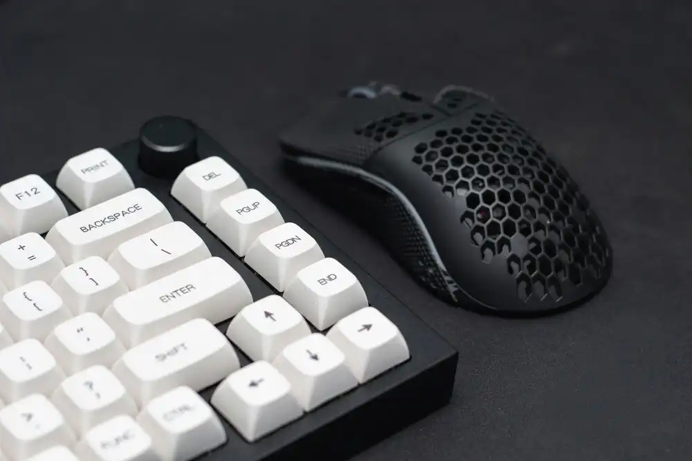 Close-up of a mechanical keyboard with white keycaps and a black gaming mouse with a hexagonal honeycomb pattern, placed on a dark surface.