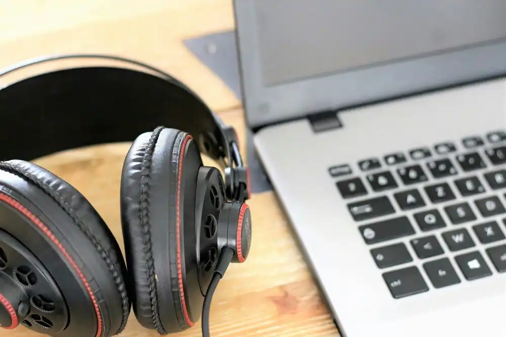 Close-up of black over-ear headphones with red stitching placed next to an open laptop on a wooden surface. The laptop keyboard is partially visible, suggesting a workspace or study setup.