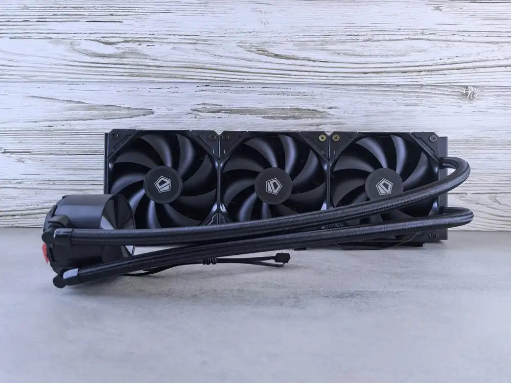 Three black cooling fans mounted in a row, attached to a liquid cooling radiator. The unit has a sleek design and rests against a light wooden background. A coiled tube extends from the side, connected to a pump with a red accent.