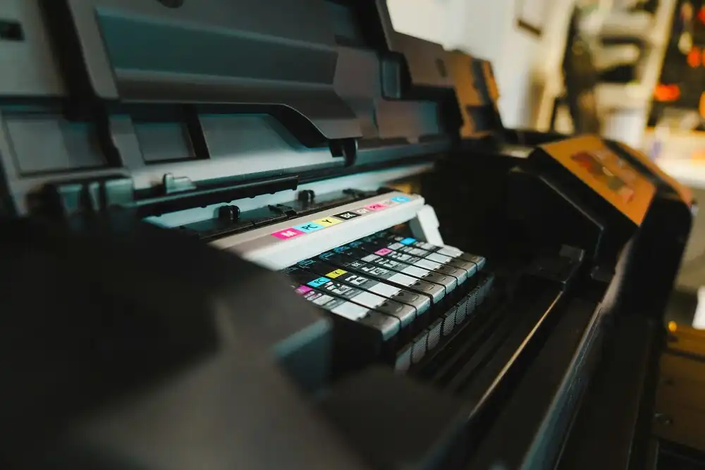 Close-up of an open printer showing a row of multi-colored ink cartridges inside. The printer is positioned in a dimly lit room, highlighting the cyan, magenta, yellow, and black (CMYK) labels on the cartridges.