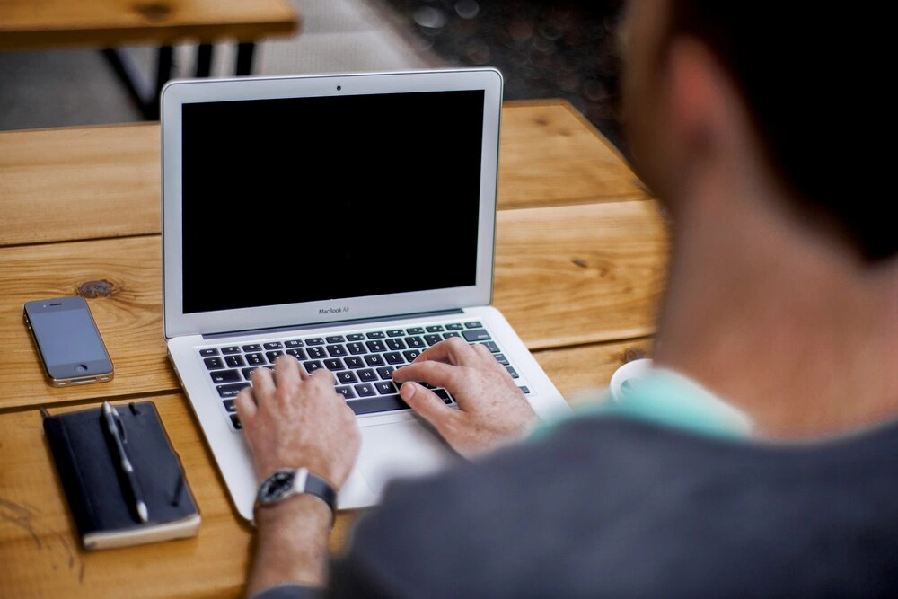 A person typing on a silver laptop at an outdoor wooden table, with a smartphone, a closed black notebook, and a white coffee cup nearby. The person is wearing a watch and casual clothing.