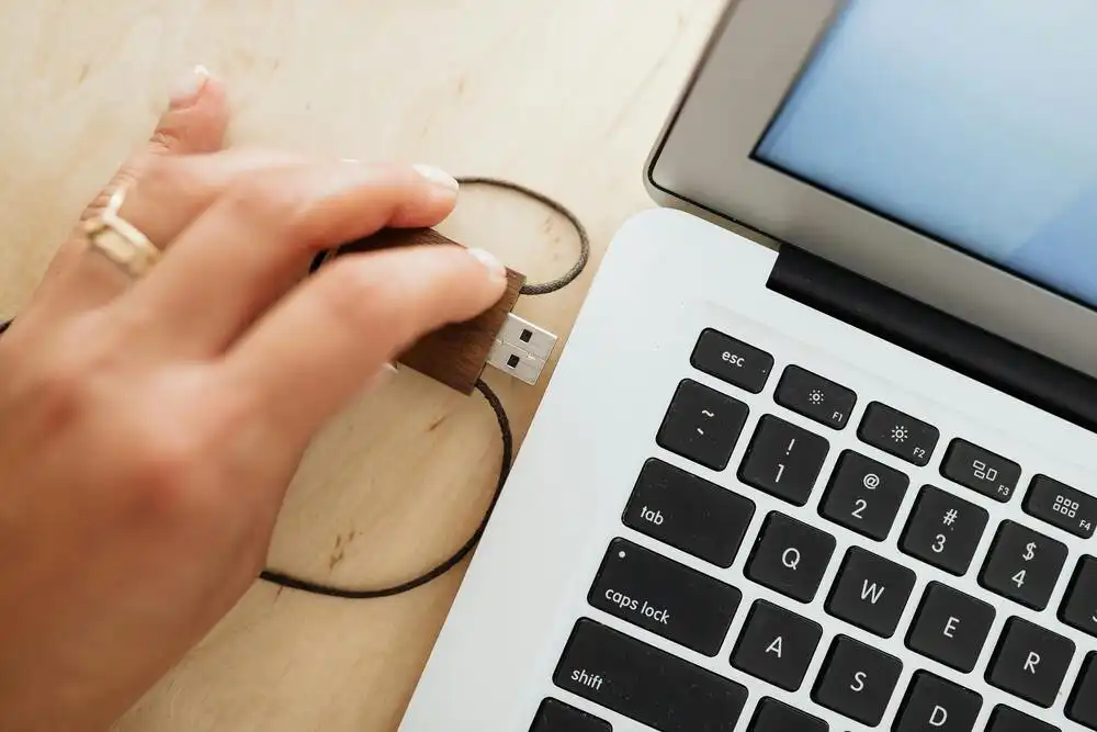 A hand plugs a USB flash drive into a laptop. The laptop is open, displaying part of the screen and keyboard on a wooden surface. The flash drive has a wooden exterior and is connected by a cord.