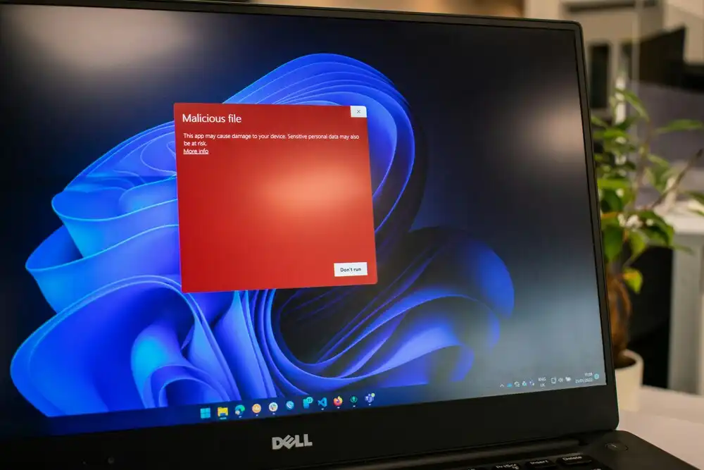 A Dell laptop screen displays a red warning pop-up labeled "Malicious file," indicating potential damage or data theft. The desktop background is the Windows 11 default wallpaper. A plant is partially visible in the background.